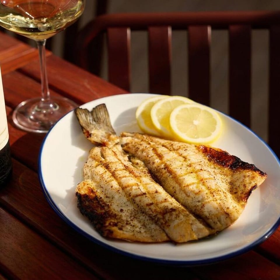 Grilled fish fillets with lemon slices on a blue-rimmed plate beside a bottle of wine and filled wineglass.