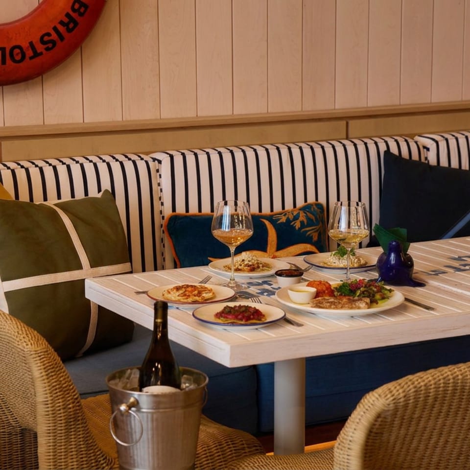 Coastal restaurant dining booth with striped upholstery, colorful pillows, seafood dishes, wine glasses, and a chilled bottle.