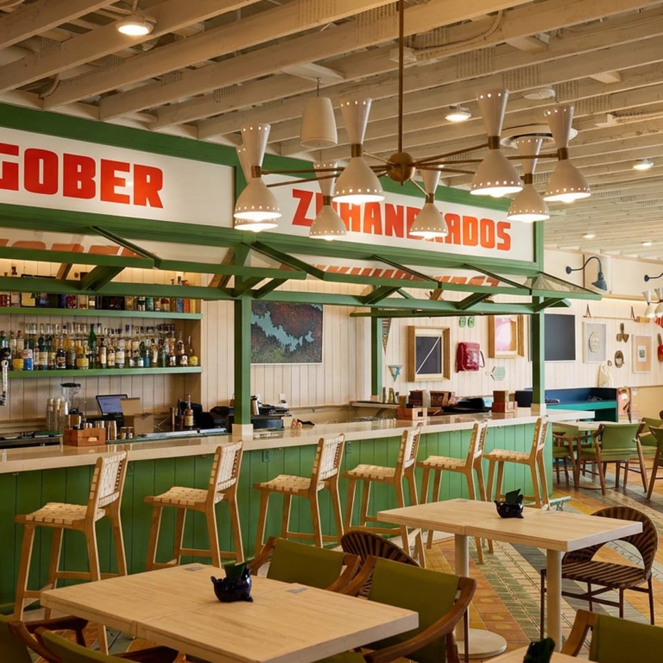 Bright restaurant interior with green bar, liquor shelves, patterned flooring, wooden tables, and woven chairs beneath decorative pendant lights.