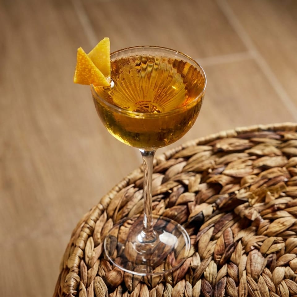 Amber cocktail in a stemmed glass garnished with orange peel, resting on a woven tabletop beside a wooden floor.