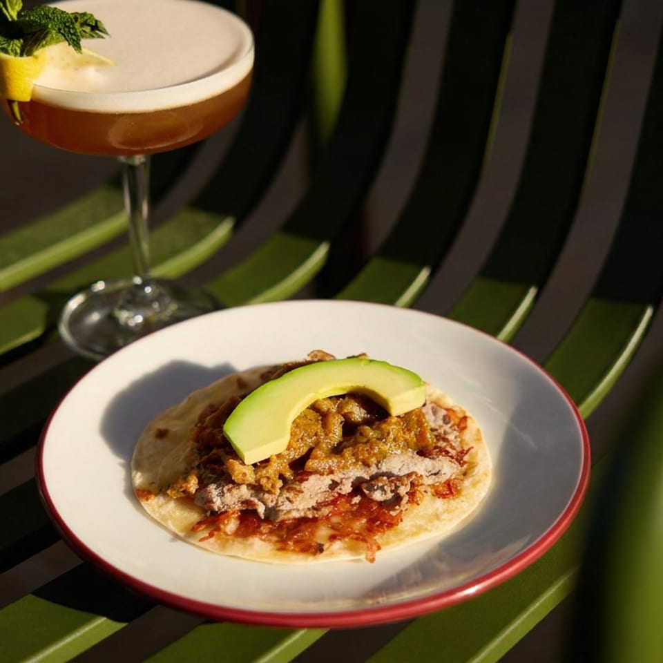Taco topped with avocado on a red-rimmed plate beside a mint-garnished cocktail on a green-and-black chair.