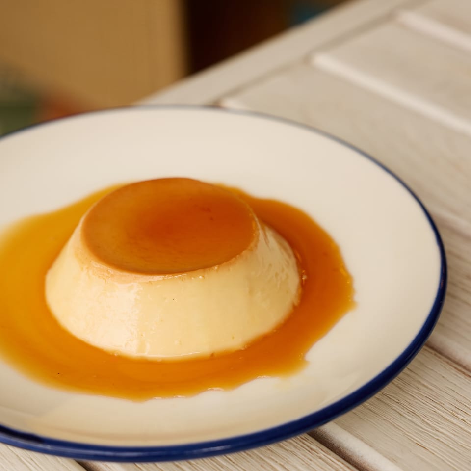 Caramel flan served on a white plate with blue trim, surrounded by glossy caramel sauce on a wooden table.