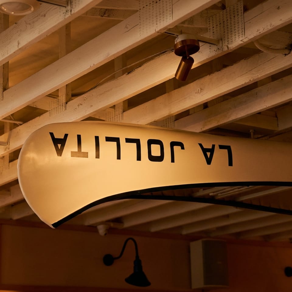 Warmly lit restaurant interior featuring a curved illuminated sign, exposed wooden beams, pendant lights, and wall sconces.