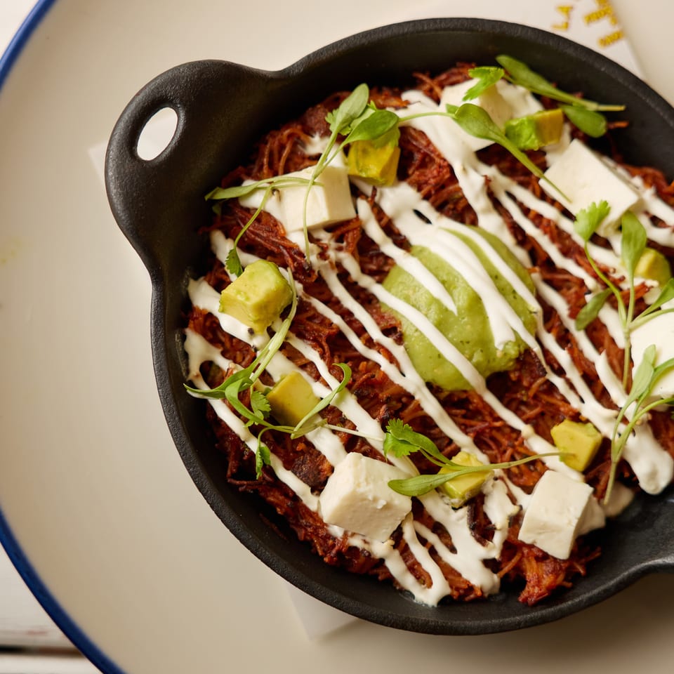 Crispy shredded dish in a black skillet, topped with creamy sauce, avocado, cheese cubes, and fresh herbs.